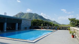 A serene outdoor swimming pool surrounded by potted plants and lounge chairs, with a backdrop of lush green hills and a blue sky.
