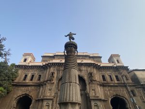 Majestic stone building with arched doorways, twin towers, and a statue-topped pillar, set against a clear blue sky.
