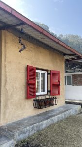 A small, rustic house with a beige clay exterior features a large white window framed by bright red wooden shutters, which are open.