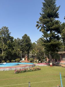 A botanical garden area behind a wire fence. The grass is cut short.  There's a line of flowers in the middle of the lawn.  There appears to be a large dry fountain in the area.