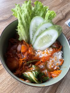
A green bowl filled with rice topped with a colorful stir-fry mix of vegetables, including cooked onions, bell peppers, and greens, garnished with fresh cucumber slices and leafy romaine lettuce.